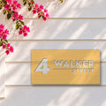 Brushed gold house number and street name sign with a brushed metal look, The Alexandra, reading 4 Walker Street in white lettering, on white weatherboards under a pink bougainvillea.