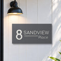 Matte charcoal house number and street name sign, The Alexandra, reading 8 Sandview Place in white lettering, on white cladding under a black wall light, beside a black front door.