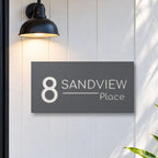Matte charcoal house number and street name sign, The Alexandra, reading 8 Sandview Place in white lettering, on white cladding under a black wall light, beside a black front door.