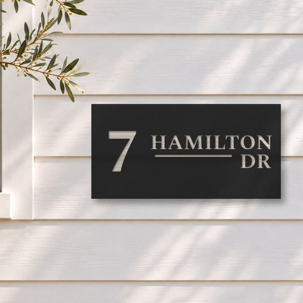 Brushed silver house number and street name sign with a brushed metal look, The Alexandra, black face reading 7 Hamilton Dr in silver lettering, on white weatherboards beside a black-framed window and an olive tree.