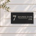 Brushed silver house number and street name sign with a brushed metal look, The Alexandra, black face reading 7 Hamilton Dr in silver lettering, on white weatherboards beside a black-framed window and an olive tree.