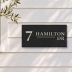 Brushed silver house number and street name sign with a brushed metal look, The Alexandra, black face reading 7 Hamilton Dr in silver lettering, on white weatherboards beside a black-framed window and an olive tree.