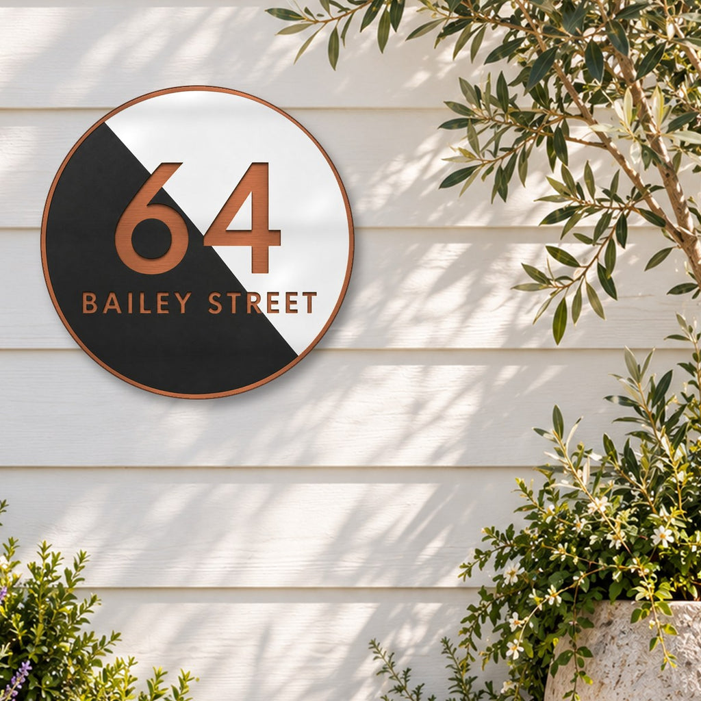 Copper round house number and street name sign reading 64 Bailey Street, split diagonally into black and white, with brushed copper lettering and a brushed copper rim, The Birtinya style, on white weatherboards beside an olive tree and flowering shrubs.