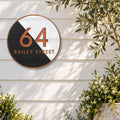 Copper round house number and street name sign reading 64 Bailey Street, split diagonally into black and white, with brushed copper lettering and a brushed copper rim, The Birtinya style, on white weatherboards beside an olive tree and flowering shrubs.