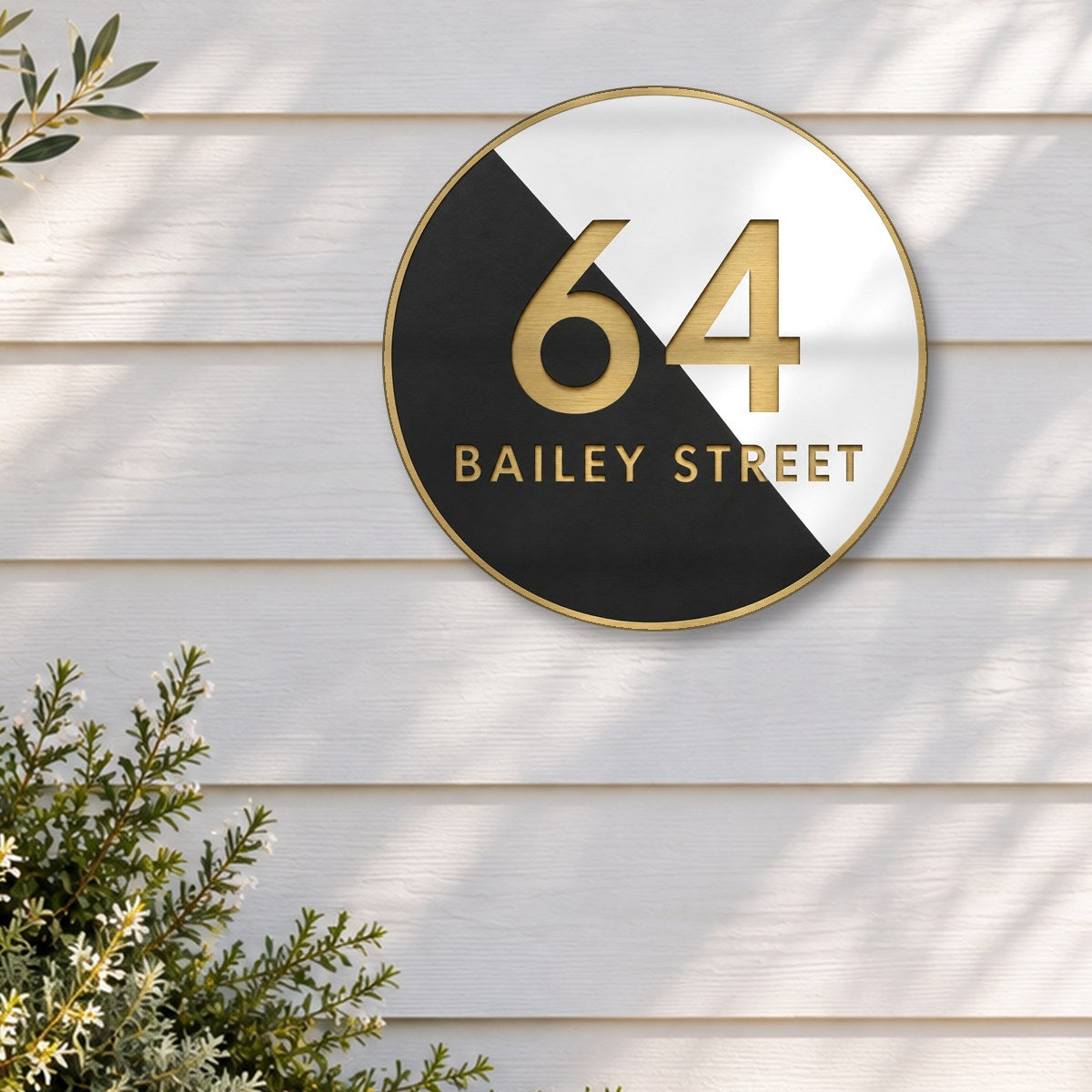 Gold round house number and street name sign reading 64 Bailey Street, split diagonally into black and white, with brushed gold lettering and a brushed gold rim, The Birtinya style, on white weatherboards above an olive tree in a stone pot.