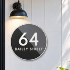 Matte round house number and street name sign reading 64 Bailey Street, split diagonally into matte black and charcoal, with white lettering and a white rim, The Birtinya style, on white cladding under a black wall light, beside a black front door.