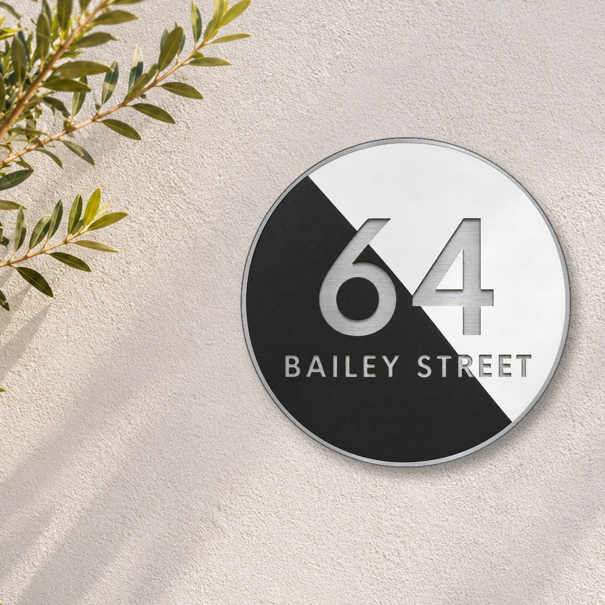 Silver round house number and street name sign reading 64 Bailey Street, split diagonally into black and white, with brushed silver lettering and a brushed silver rim, The Birtinya style, on a white rendered wall with olive branches.