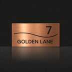 Brushed copper house number and street name sign with a brushed metal look, The Bokarina, reading 7 Golden Lane in black lettering, with a wave line.