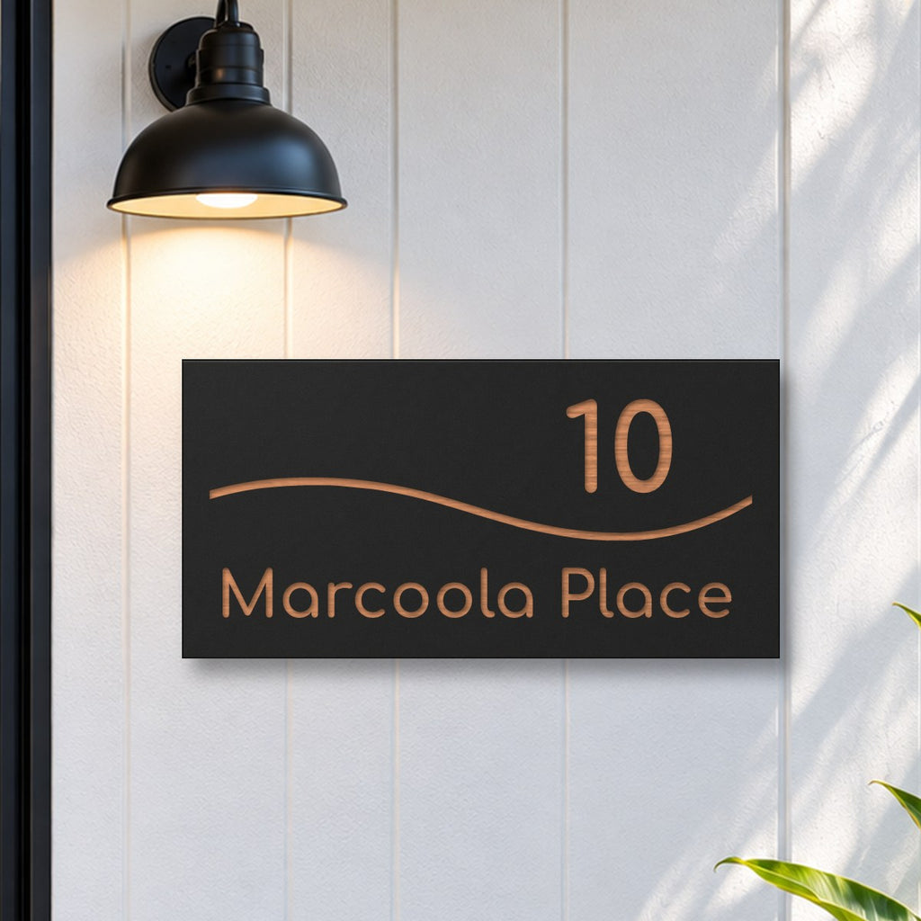 Brushed copper house number and street name sign with a brushed metal look, The Bokarina, black face reading 10 Marcoola Place in copper lettering, with a wave line, on white cladding under a black wall light, beside a black front door.
