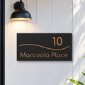 Brushed copper house number and street name sign with a brushed metal look, The Bokarina, black face reading 10 Marcoola Place in copper lettering, with a wave line, on white cladding under a black wall light, beside a black front door.