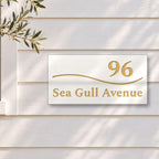 Brushed gold house number and street name sign with a brushed metal look, The Bokarina, white face reading 96 Sea Gull Avenue in gold lettering, with a wave line, on white weatherboards beside a black-framed window and an olive tree.