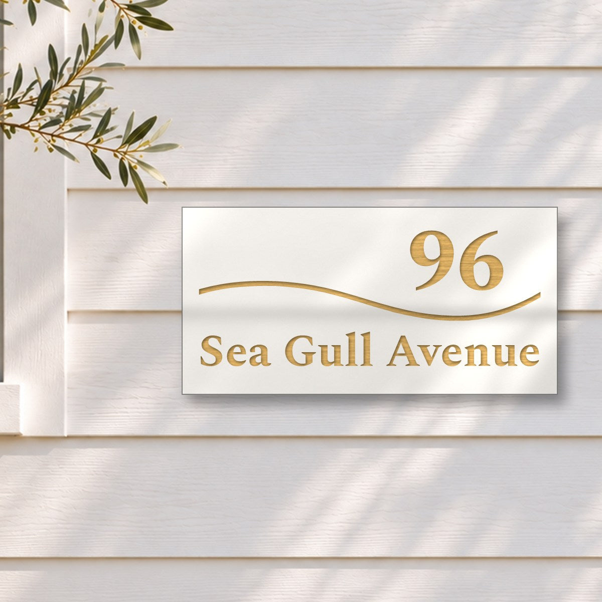 Brushed gold house number and street name sign with a brushed metal look, The Bokarina, white face reading 96 Sea Gull Avenue in gold lettering, with a wave line, on white weatherboards beside a black-framed window and an olive tree.