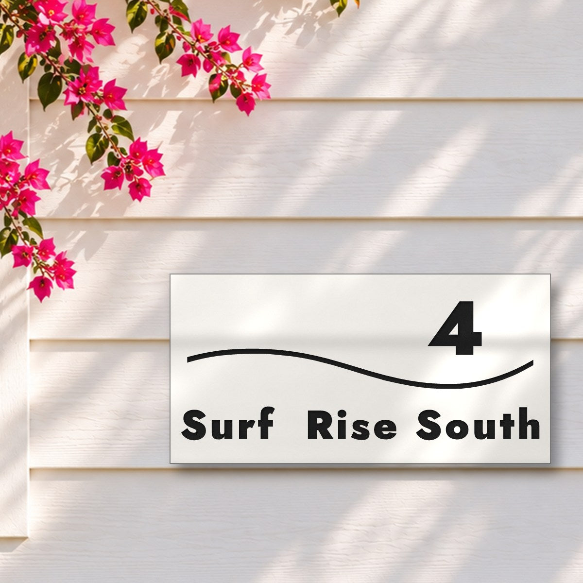 Matte white house number and street name sign, The Bokarina, reading 4 Surf Rise South in black lettering, with a wave line, on white weatherboards under a pink bougainvillea.