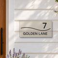 Brushed silver house number and street name sign with a brushed metal look, The Bokarina, reading 7 Golden Lane in black lettering, with a wave line, on white weatherboards beside a timber front door, with lavender and an olive tree.