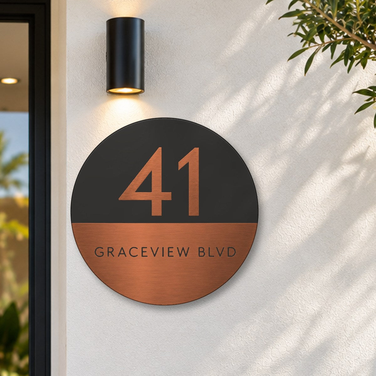 Copper round house number and street name sign reading 41 Graceview Blvd, with a black top half and a copper number, over a brushed copper bottom half and a black street name, The Buddina style, on a white rendered wall under a black wall light, beside the front door.