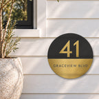 Gold round house number and street name sign reading 41 Graceview Blvd, with a black top half and a gold number, over a brushed gold bottom half and a black street name, The Buddina style, on white weatherboards below a black-framed window, beside an olive tree.