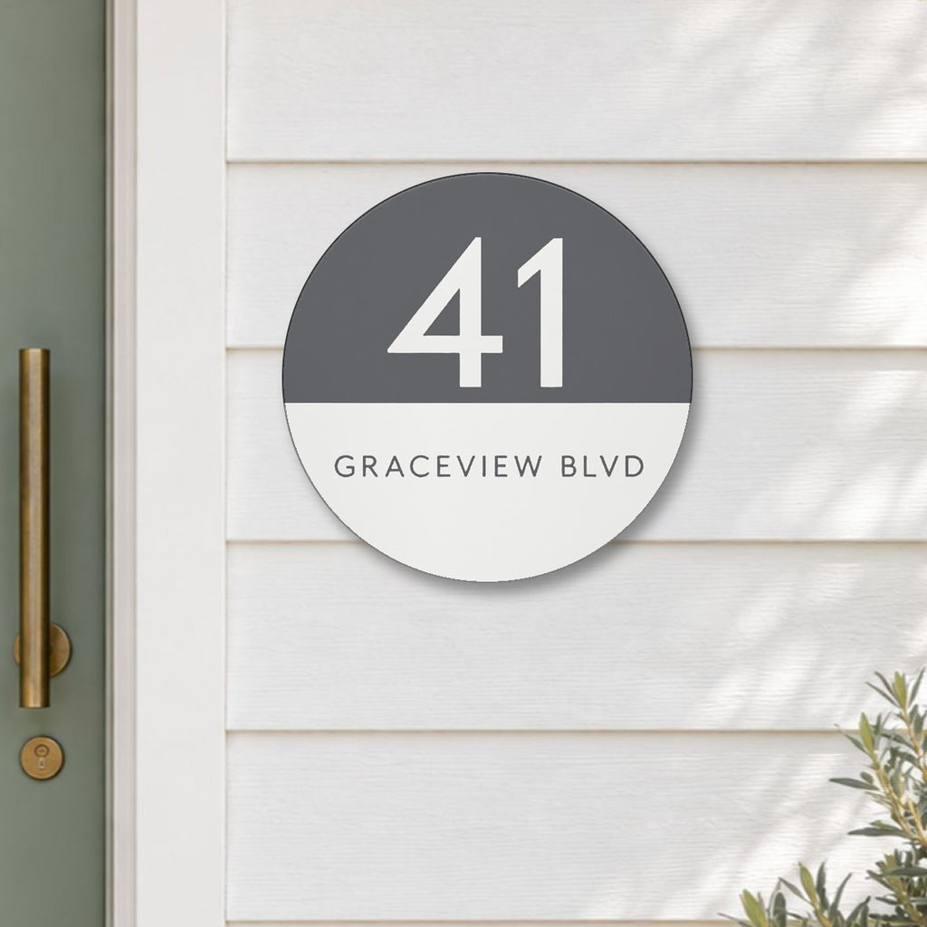 Matte round house number and street name sign reading 41 Graceview Blvd, with a matte charcoal top half and a white number, over a white bottom half and a charcoal street name, The Buddina style, on white weatherboards beside a sage green front door.