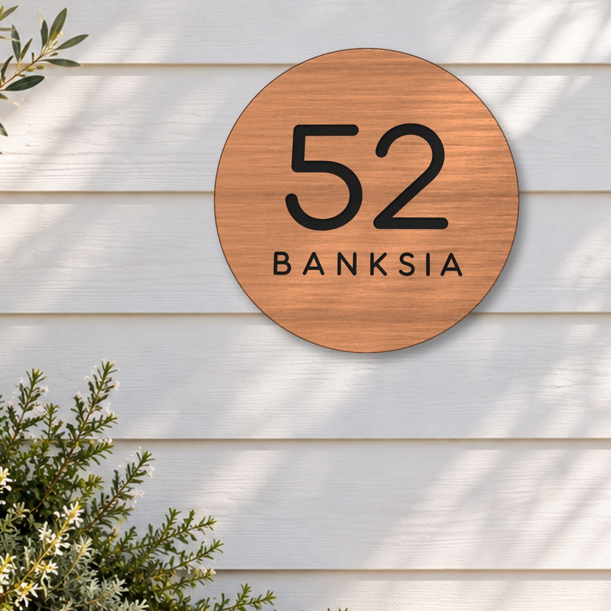 Brushed copper round house number and street name sign reading 52 Banksia cut through to black, The Buderim style, on white weatherboards above an olive tree in a stone pot.