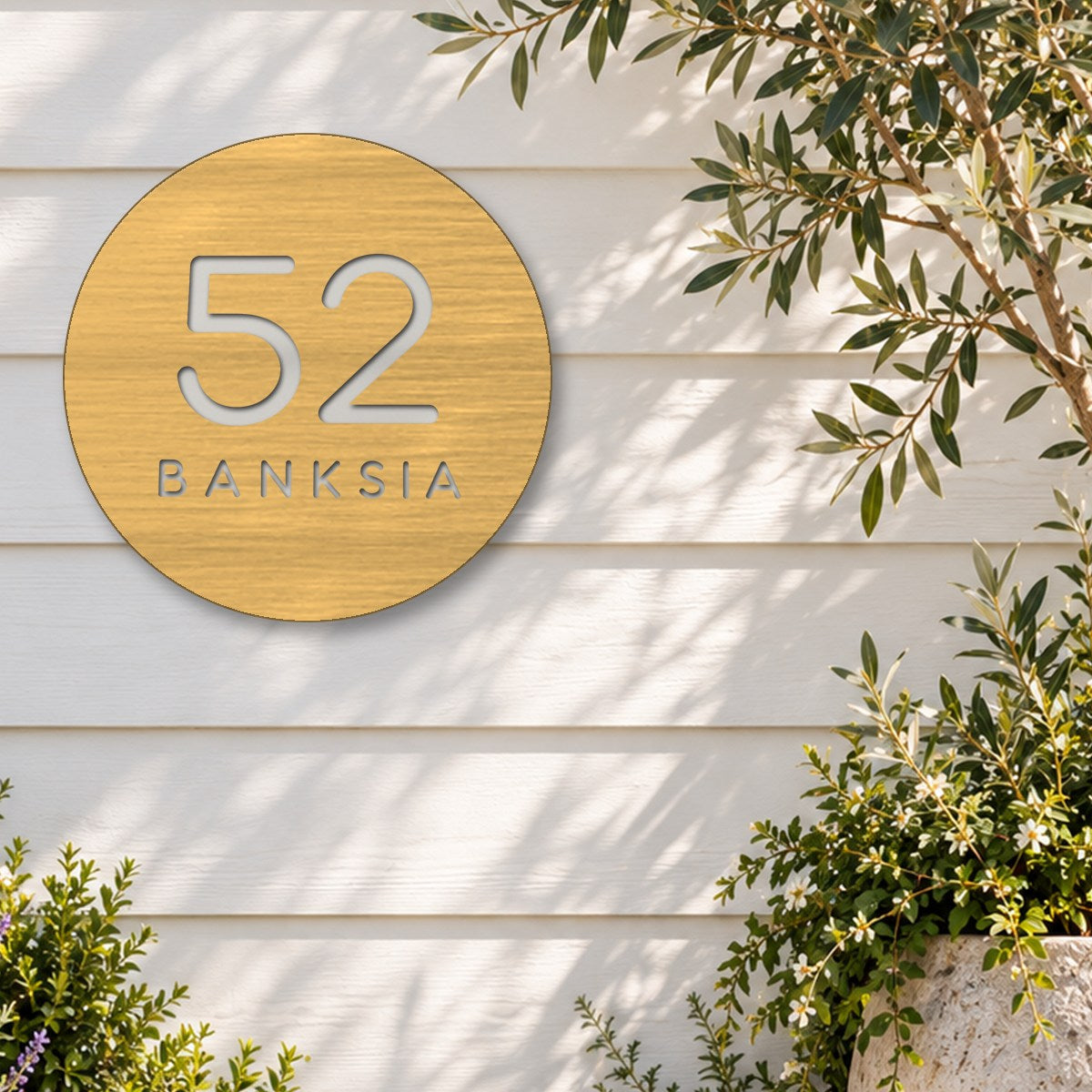 Brushed gold round house number and street name sign reading 52 Banksia cut through to white, The Buderim style, on white weatherboards beside an olive tree and flowering shrubs.