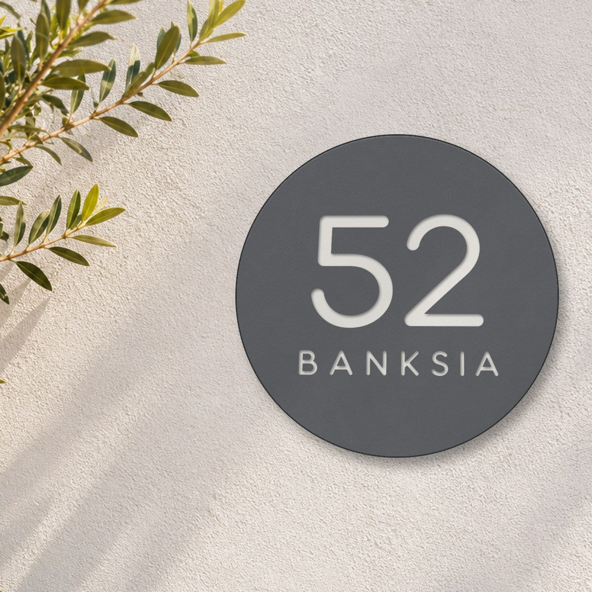 Matte charcoal round house number and street name sign reading 52 Banksia cut through to white, The Buderim style, on a white rendered wall with olive branches.