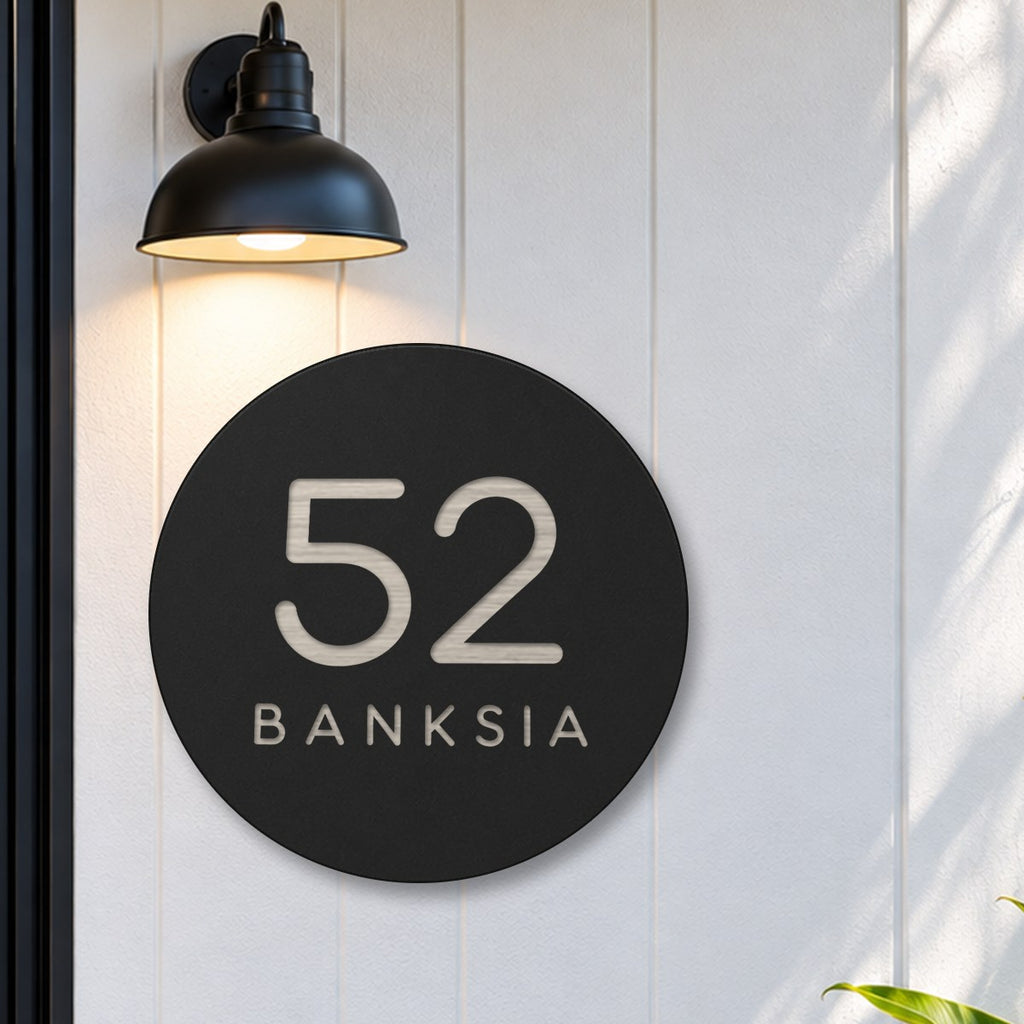 Black round house number and street name sign reading 52 Banksia cut through to brushed silver, The Buderim style, on white cladding under a black wall light, beside a black front door.