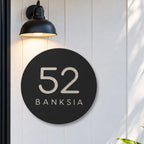 Black round house number and street name sign reading 52 Banksia cut through to brushed silver, The Buderim style, on white cladding under a black wall light, beside a black front door.