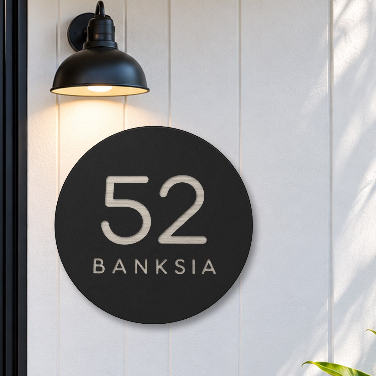 Black round house number and street name sign reading 52 Banksia cut through to brushed silver, The Buderim style, on white cladding under a black wall light, beside a black front door.