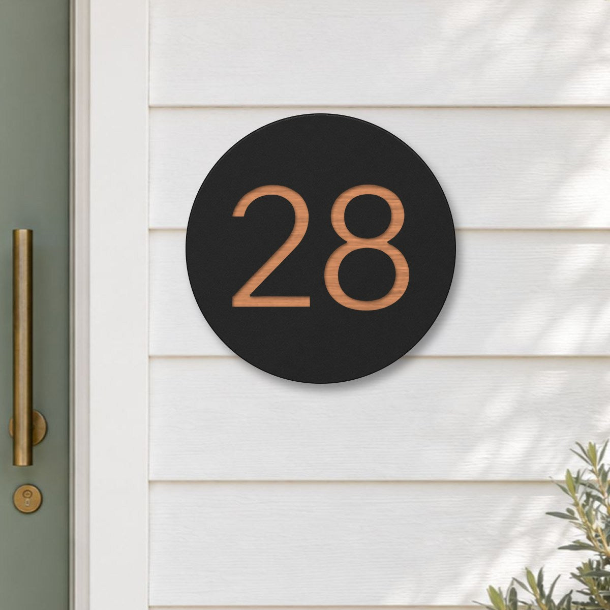 Black round house number sign with the number 28 cut through to brushed copper, The Caloundra style, on white weatherboards beside a sage green front door.