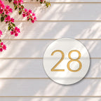 White round house number sign with the number 28 cut through to brushed gold, The Caloundra style, on white weatherboards under a pink bougainvillea.