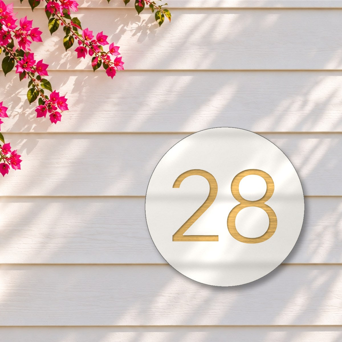 White round house number sign with the number 28 cut through to brushed gold, The Caloundra style, on white weatherboards under a pink bougainvillea.