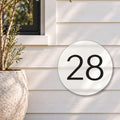 Matte white round house number sign with the number 28 cut through to black, The Caloundra style, on white weatherboards below a black-framed window, beside an olive tree.