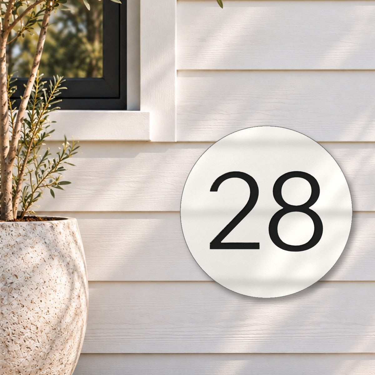 Matte white round house number sign with the number 28 cut through to black, The Caloundra style, on white weatherboards below a black-framed window, beside an olive tree.