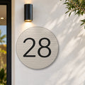 Brushed silver round house number sign with the number 28 cut through to black, The Caloundra style, on a white rendered wall under a black wall light, beside the front door.
