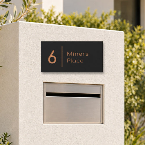 Brushed copper house number and street name sign with a brushed metal look, The Coolum, black face reading 6 Miners Place in copper lettering, with a dividing line, on a white rendered pillar above a letterbox.