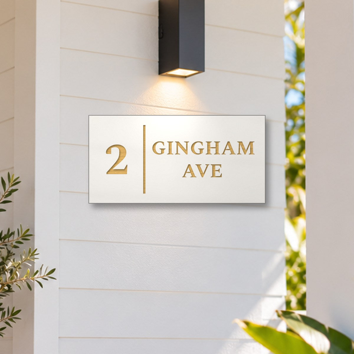 Brushed gold house number and street name sign with a brushed metal look, The Coolum, white face reading 2 Gingham Ave in gold lettering, with a dividing line, on white weatherboards under a black wall light, beside a timber front door.