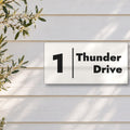 Matte white house number and street name sign, The Coolum, reading 1 Thunder Drive in black lettering, with a dividing line, on white weatherboards beside an olive tree.