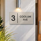 Brushed silver house number and street name sign with a brushed metal look, The Coolum, reading 3 Coolum Ave in black lettering, with a dividing line, on a white rendered wall under a wall light, beside a timber front door.