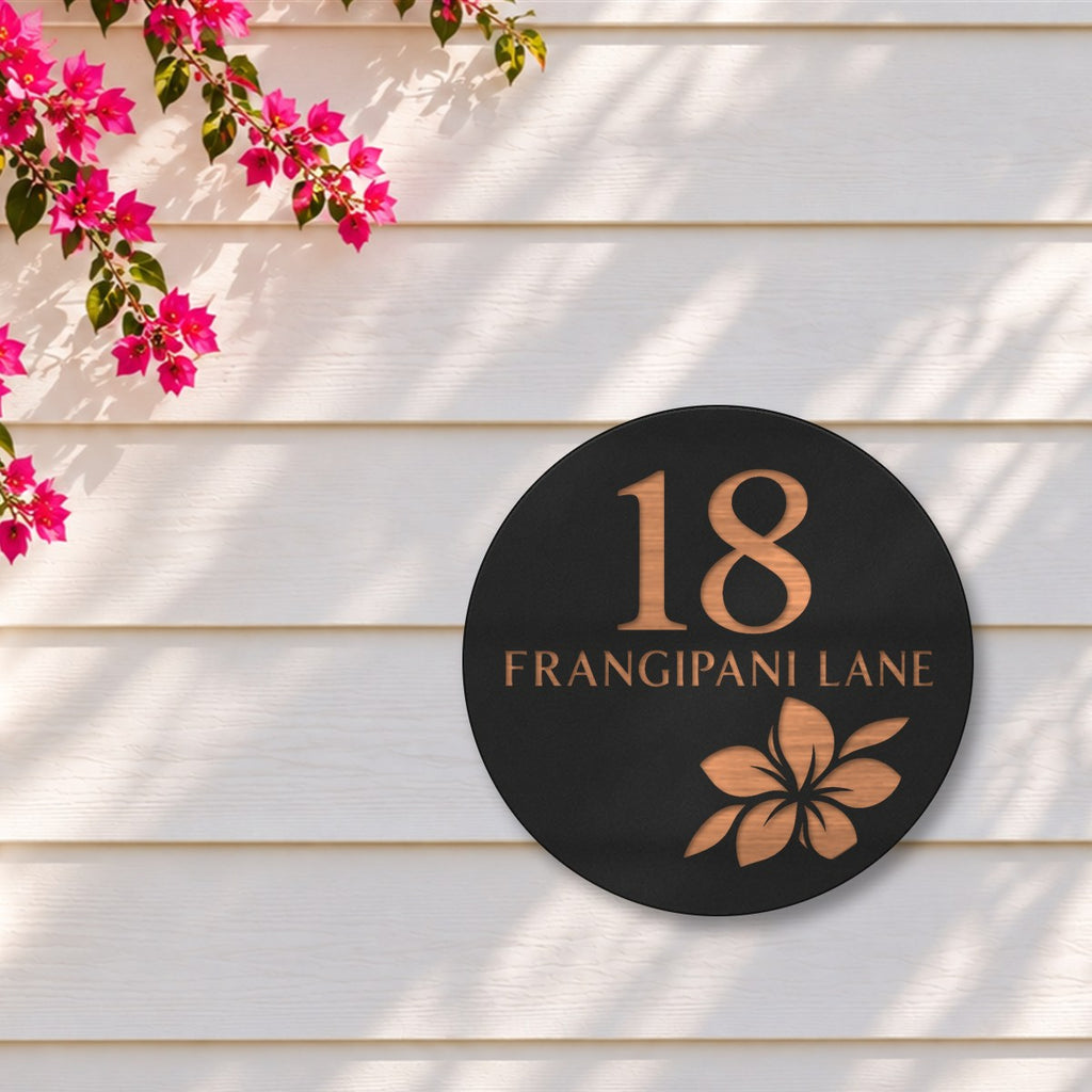 Black round house number and street name sign reading 18 Frangipani Lane with a frangipani flower cut through to brushed copper, The Currimundi style, on white weatherboards under a pink bougainvillea.