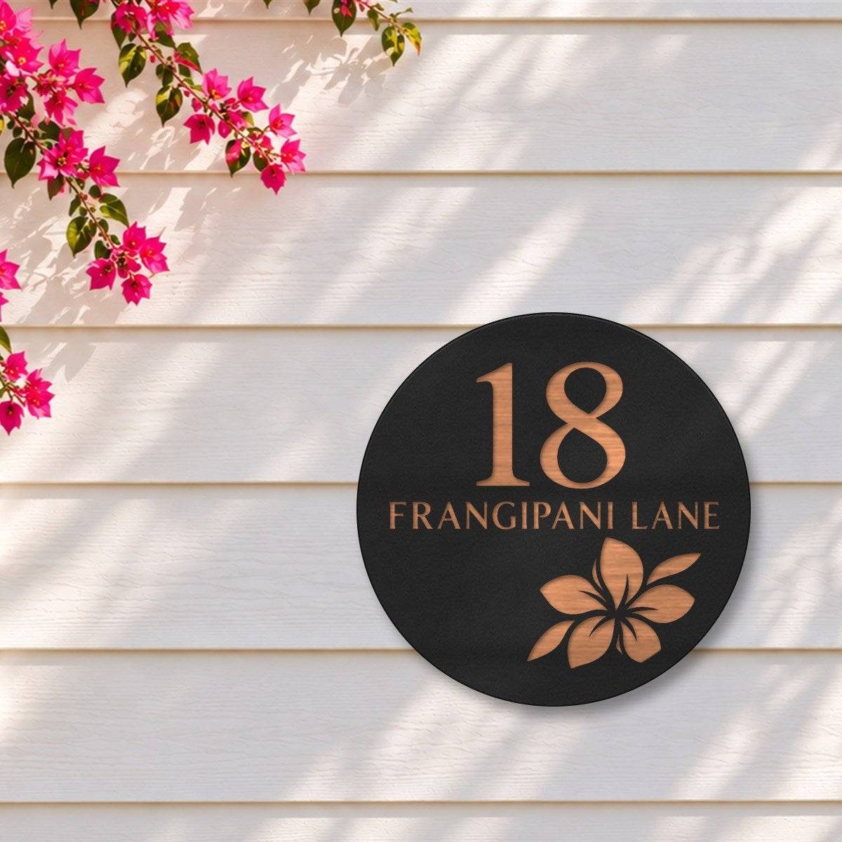 Black round house number and street name sign reading 18 Frangipani Lane with a frangipani flower cut through to brushed copper, The Currimundi style, on white weatherboards under a pink bougainvillea.