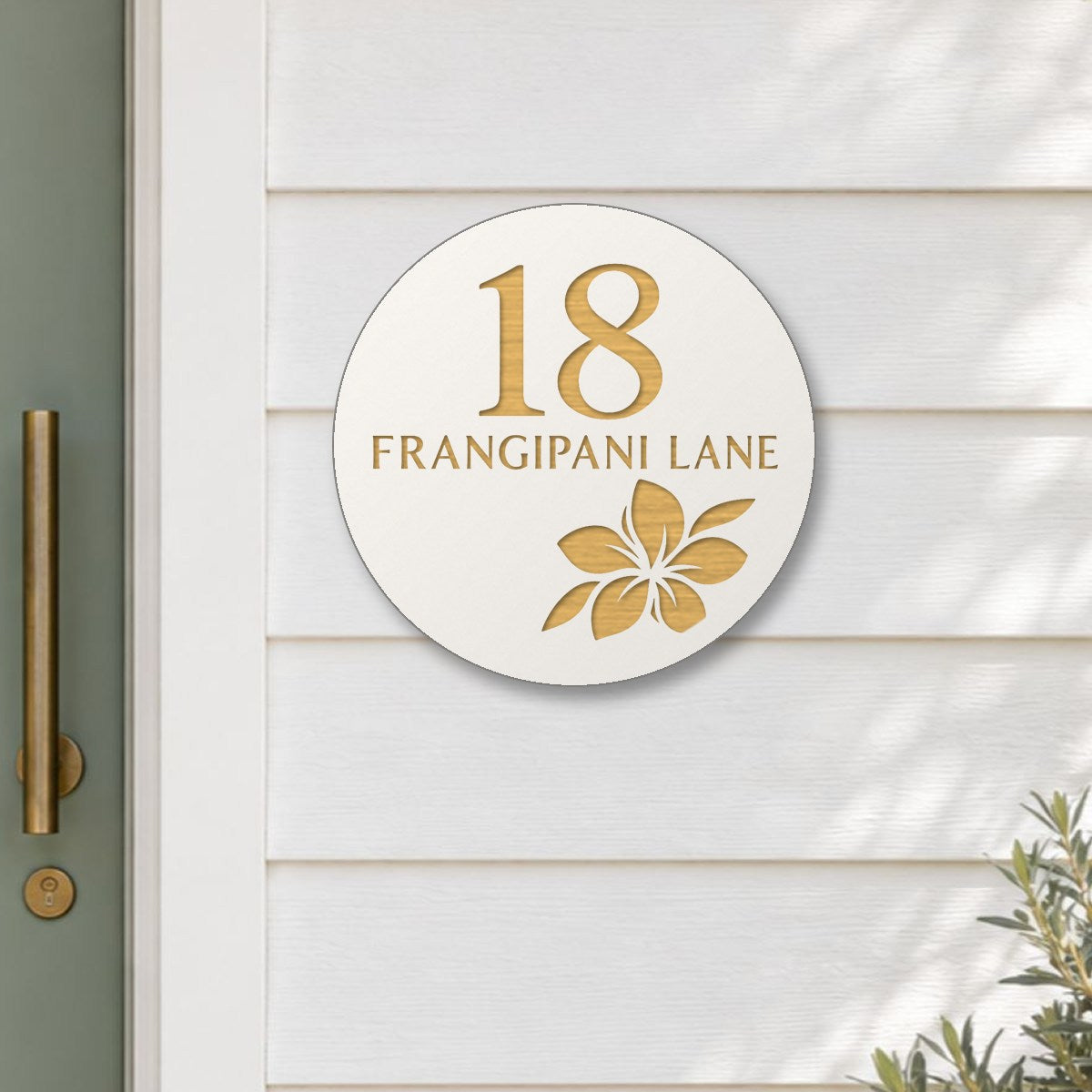 White round house number and street name sign reading 18 Frangipani Lane with a frangipani flower cut through to brushed gold, The Currimundi style, on white weatherboards beside a sage green front door.