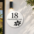 Matte white round house number and street name sign reading 18 Frangipani Lane with a frangipani flower cut through to black, The Currimundi style, on a white rendered wall under a black wall light, beside the front door.