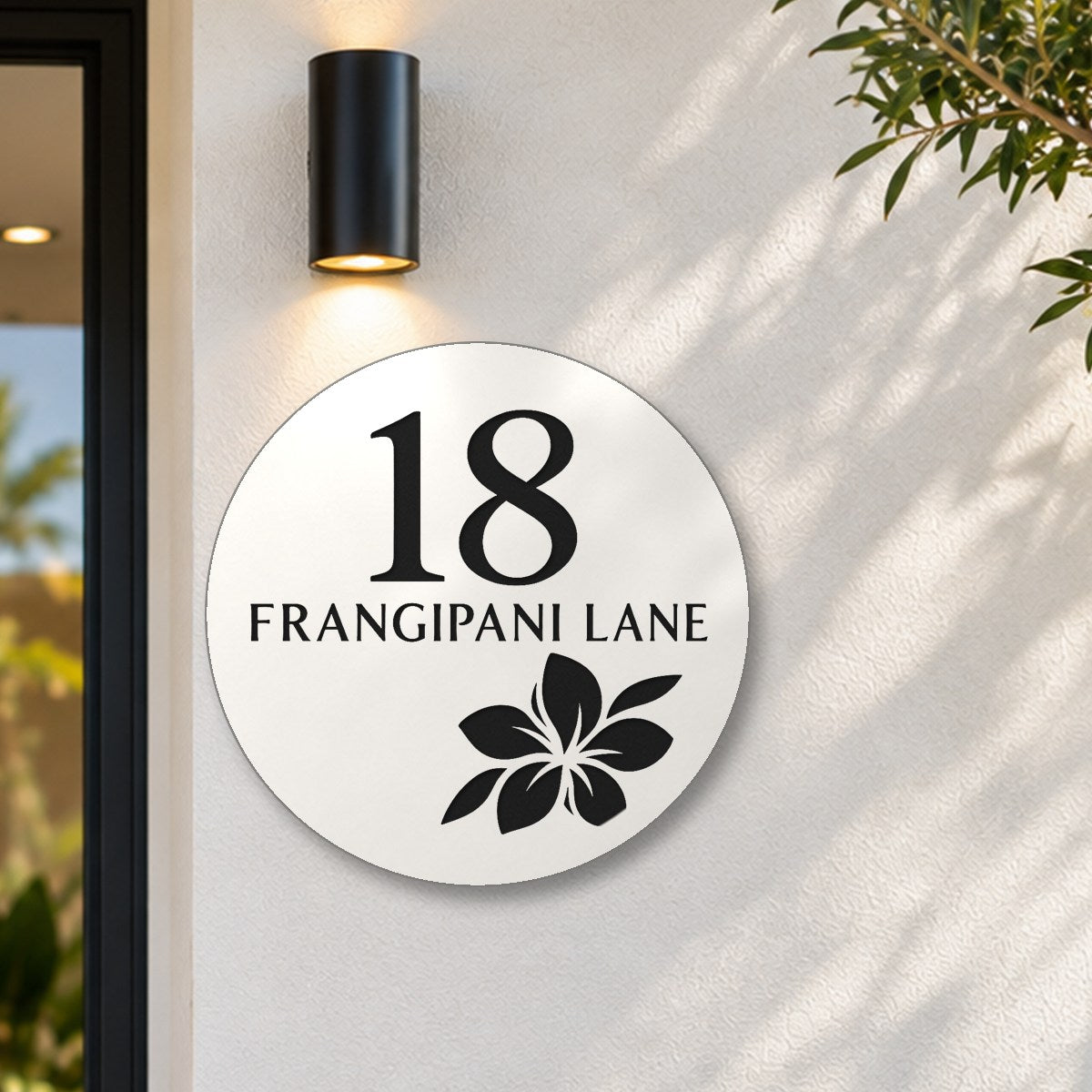 Matte white round house number and street name sign reading 18 Frangipani Lane with a frangipani flower cut through to black, The Currimundi style, on a white rendered wall under a black wall light, beside the front door.