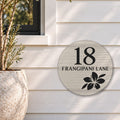 Brushed silver round house number and street name sign reading 18 Frangipani Lane with a frangipani flower cut through to black, The Currimundi style, on white weatherboards below a black-framed window, beside an olive tree.