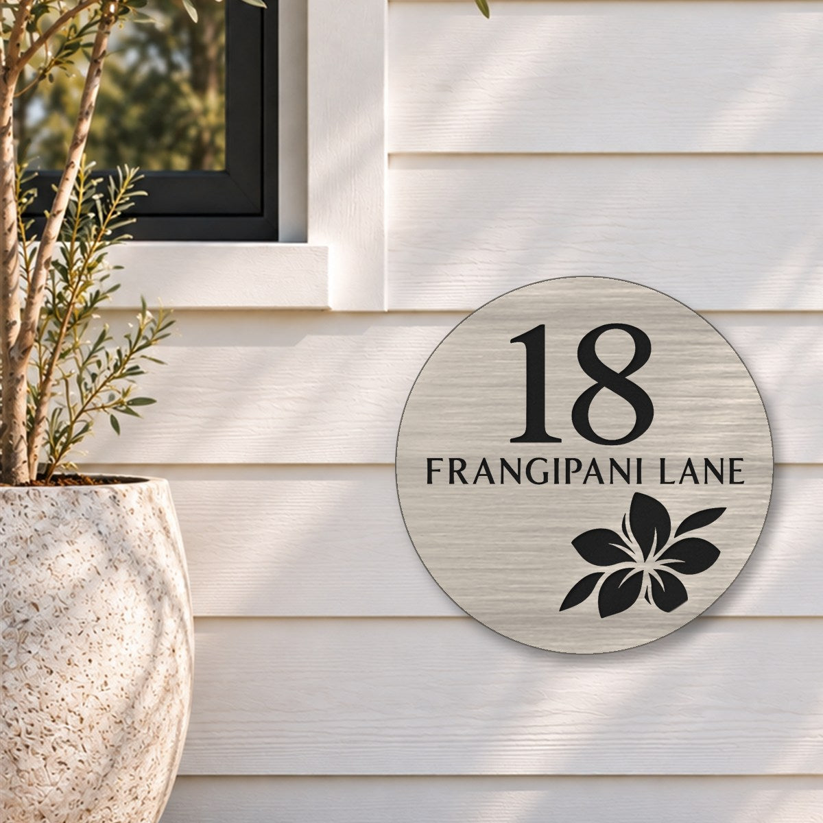 Brushed silver round house number and street name sign reading 18 Frangipani Lane with a frangipani flower cut through to black, The Currimundi style, on white weatherboards below a black-framed window, beside an olive tree.