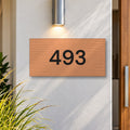 Brushed copper house number sign with a brushed metal look, The Kawana, with the number 493 in black lettering, on a white rendered wall under a wall light, beside a timber front door.