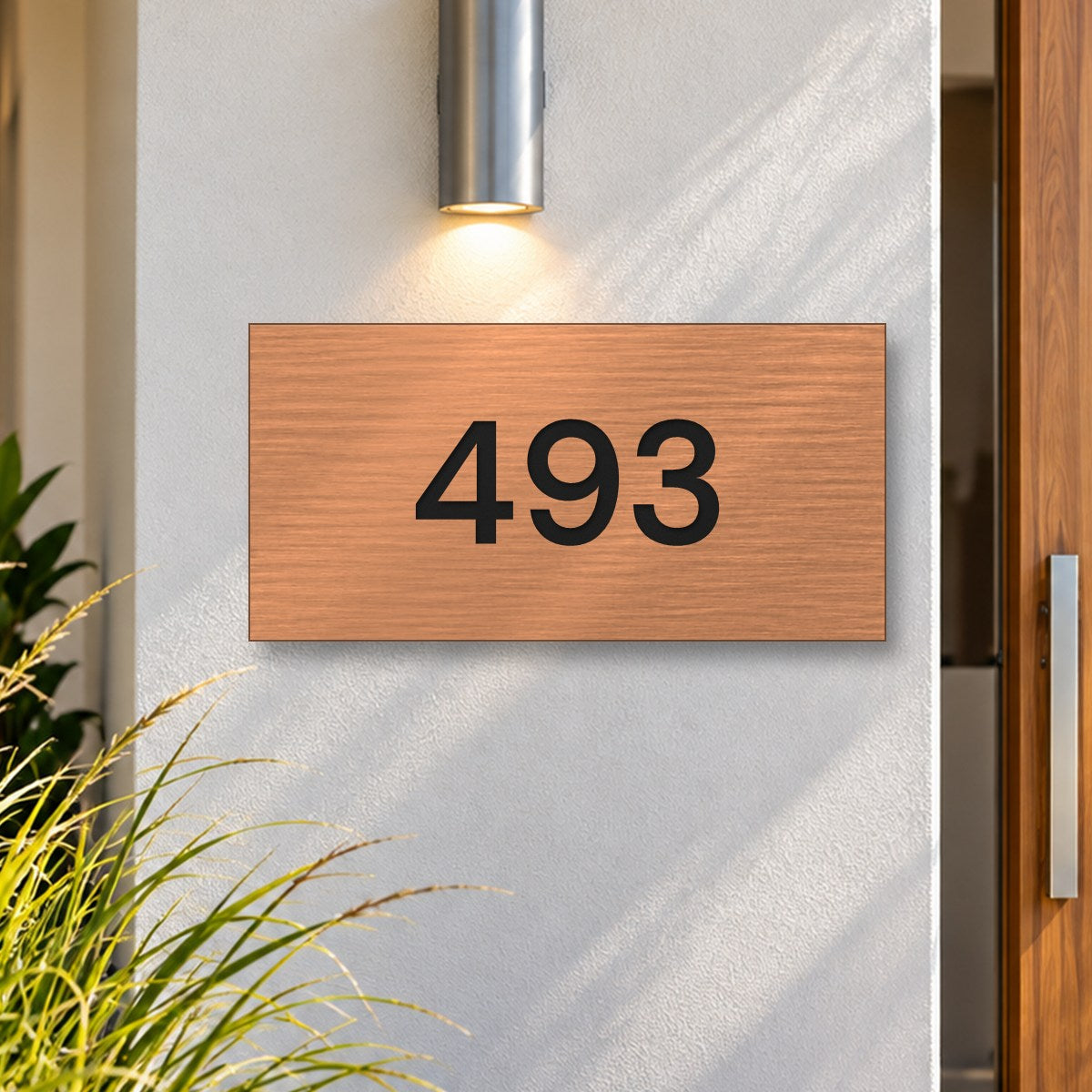 Brushed copper house number sign with a brushed metal look, The Kawana, with the number 493 in black lettering, on a white rendered wall under a wall light, beside a timber front door.