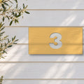Brushed gold house number sign with a brushed metal look, The Kawana, with the number 3 in white lettering, on white weatherboards beside an olive tree.