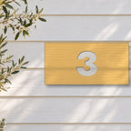 Brushed gold house number sign with a brushed metal look, The Kawana, with the number 3 in white lettering, on white weatherboards beside an olive tree.