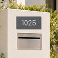 Matte charcoal house number sign, The Kawana, with the number 1025 in white lettering, on a white rendered pillar above a letterbox.