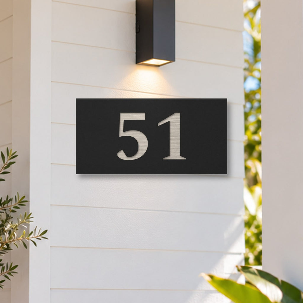 Brushed silver house number sign with a brushed metal look, The Kawana, black face with the number 51 in silver lettering, on white weatherboards under a black wall light, beside a timber front door.